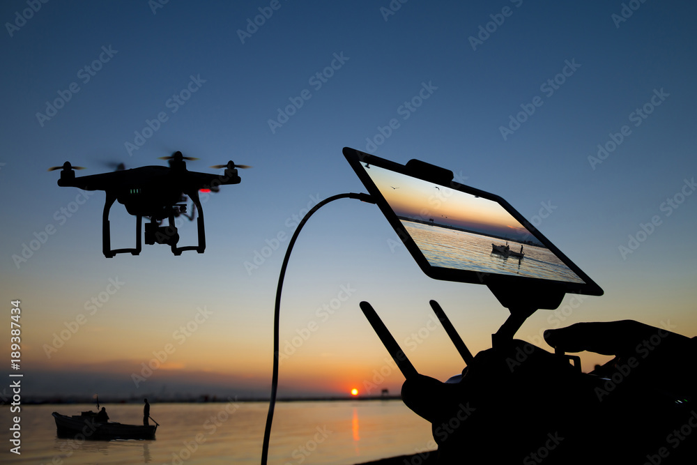A quadcopter drone hovering beside an operator's ground control tablet against a sunset over open water.