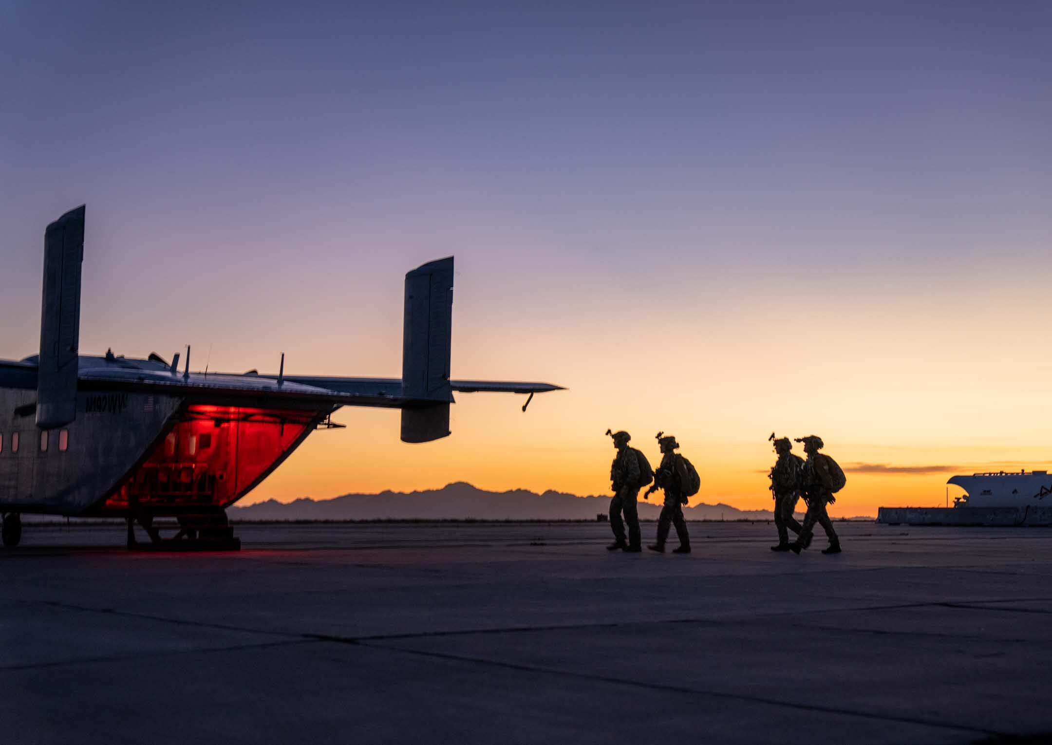 Four operators in full kit walking toward a waiting aircraft, its ramp lit red, on a desert flight line at sunset