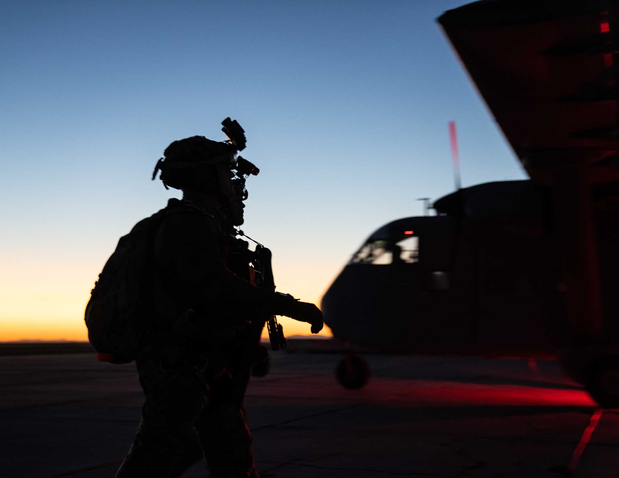 An operator in a night-vision helmet and pack silhouetted against dusk as he walks past an aircraft